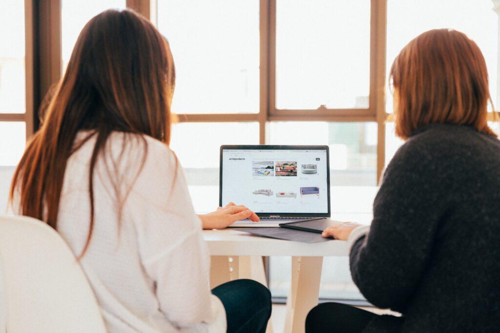 Two people sitting at a table in a bright room, looking at a laptop screen displaying a website.
