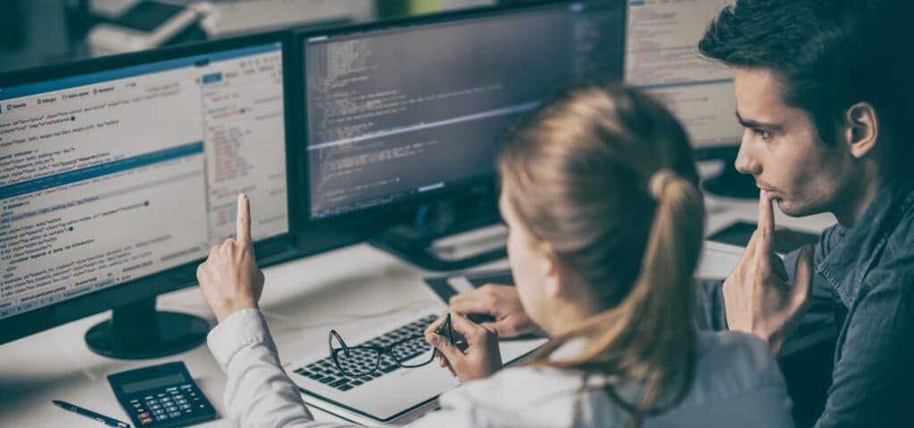 Two people at desks looking at multiple computer screens displaying code.