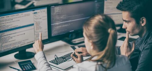 Two people at desks looking at multiple computer screens displaying code.