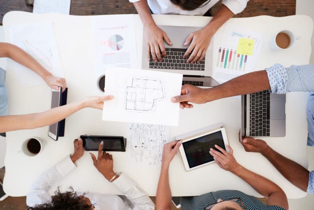 A top view of a group of people working on laptops and tablets, sharing a floor plan and charts at a conference table.