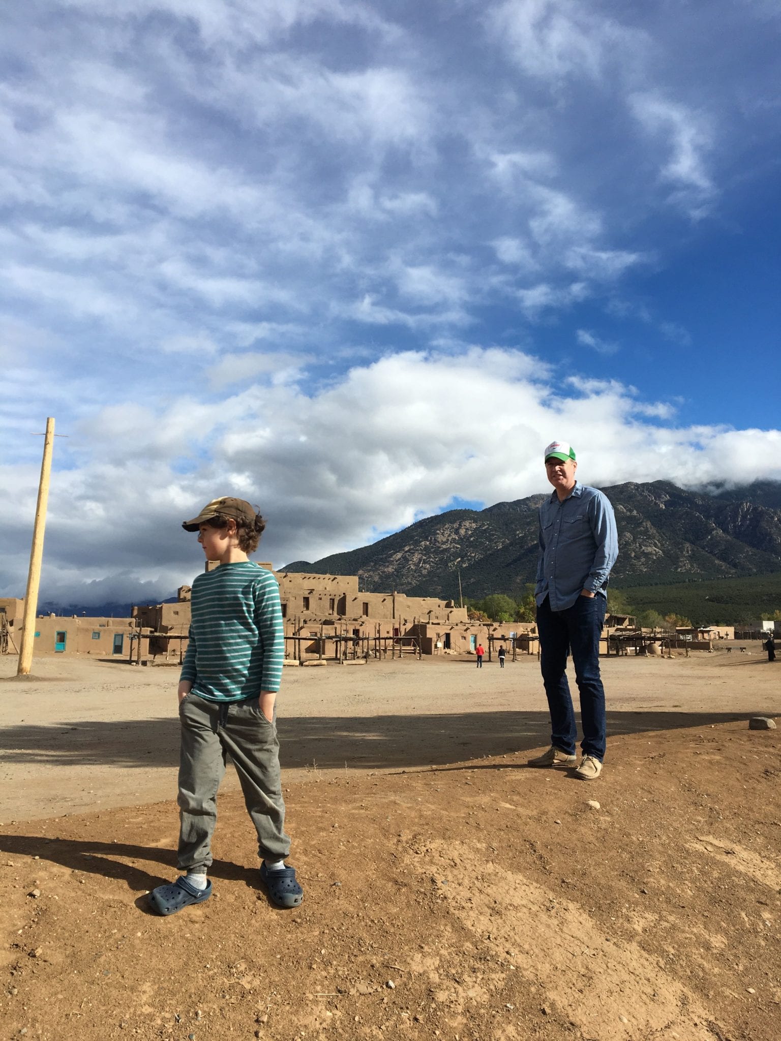 Alex Bajoris and his son standing on sandy terrain, with adobe buildings and mountains in the background under a cloudy sky.
