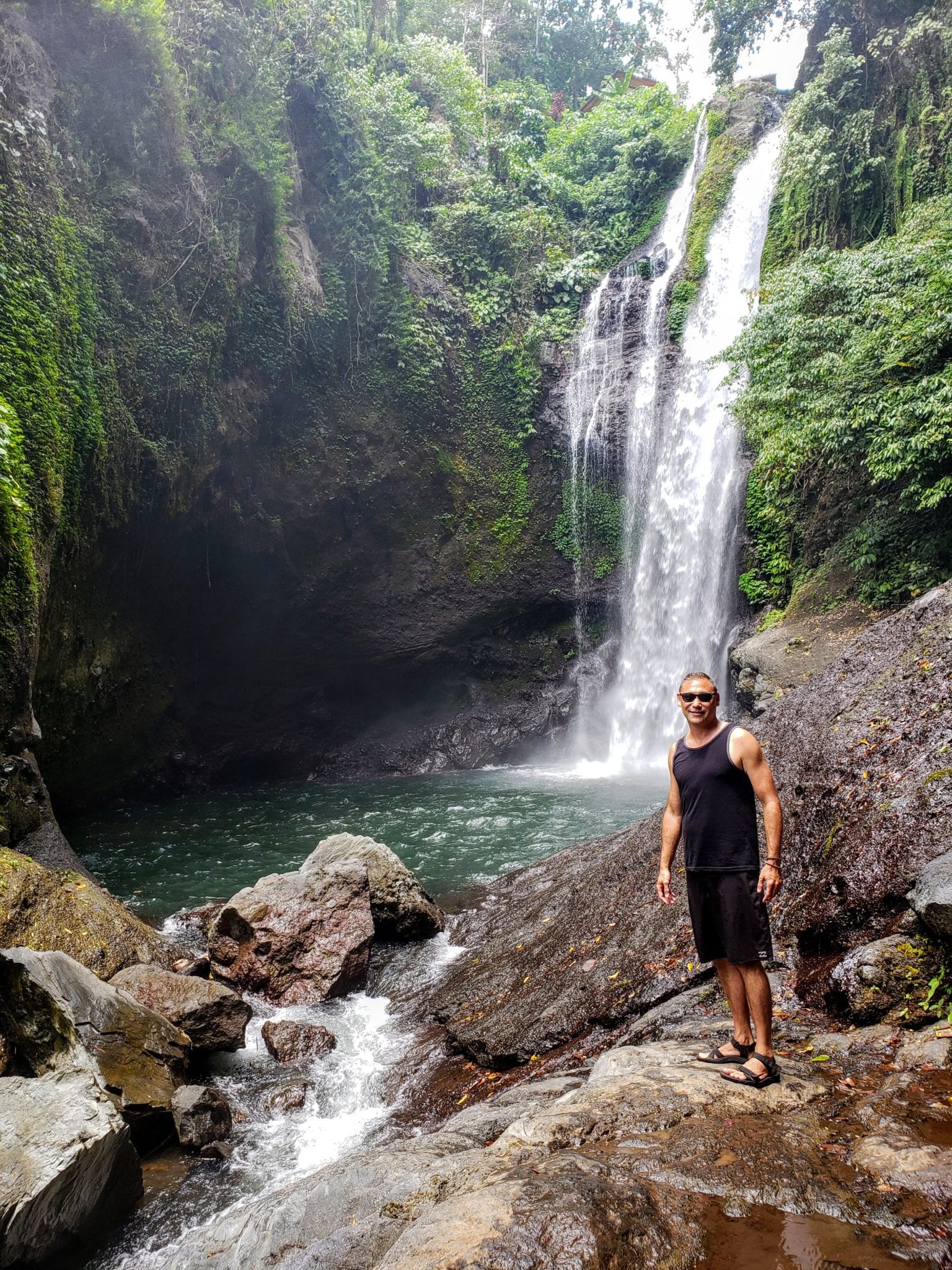 A man standing on rocks by a waterfall in Indonesia surrounded by lush greenery.