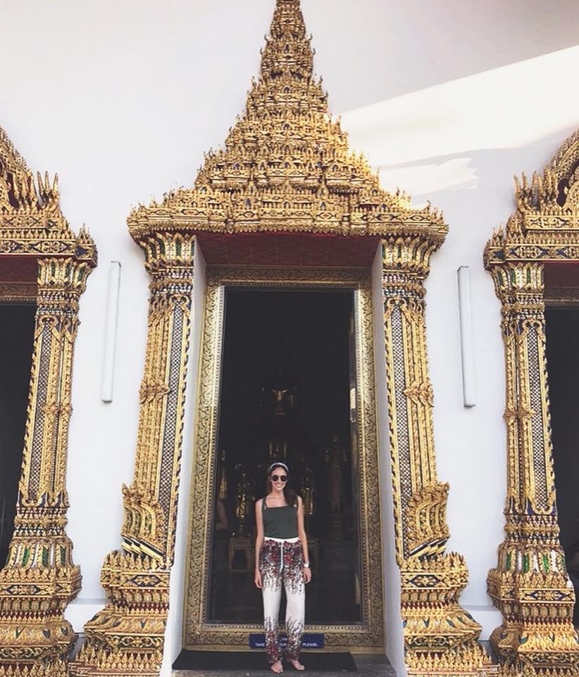 A woman standing in an ornate, gold-decorated temple doorway, wearing sunglasses and casual clothes.