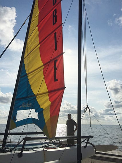Alan Bird standing on a catamaran with colorful sails, silhouetted against the ocean and cloudy sky.