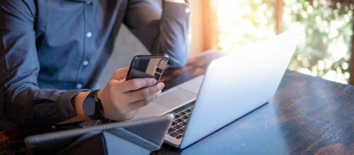 A person wearing a smartwatch using a smartphone, tablet, and laptop at a wooden table.