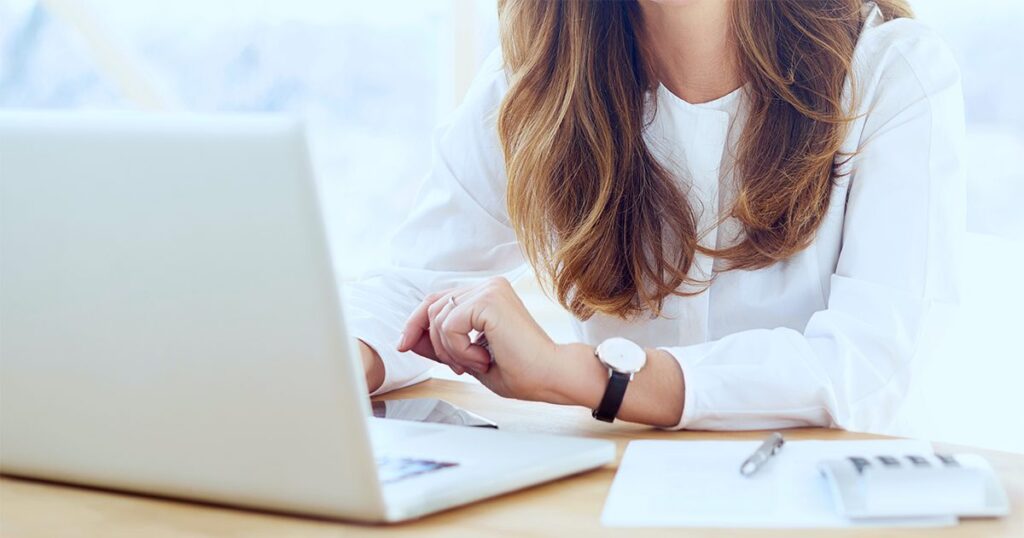 A person in a white shirt using a laptop at a desk, with a notebook and pen nearby.