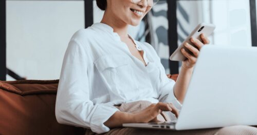 A person smiling while looking at a phone and using a laptop.