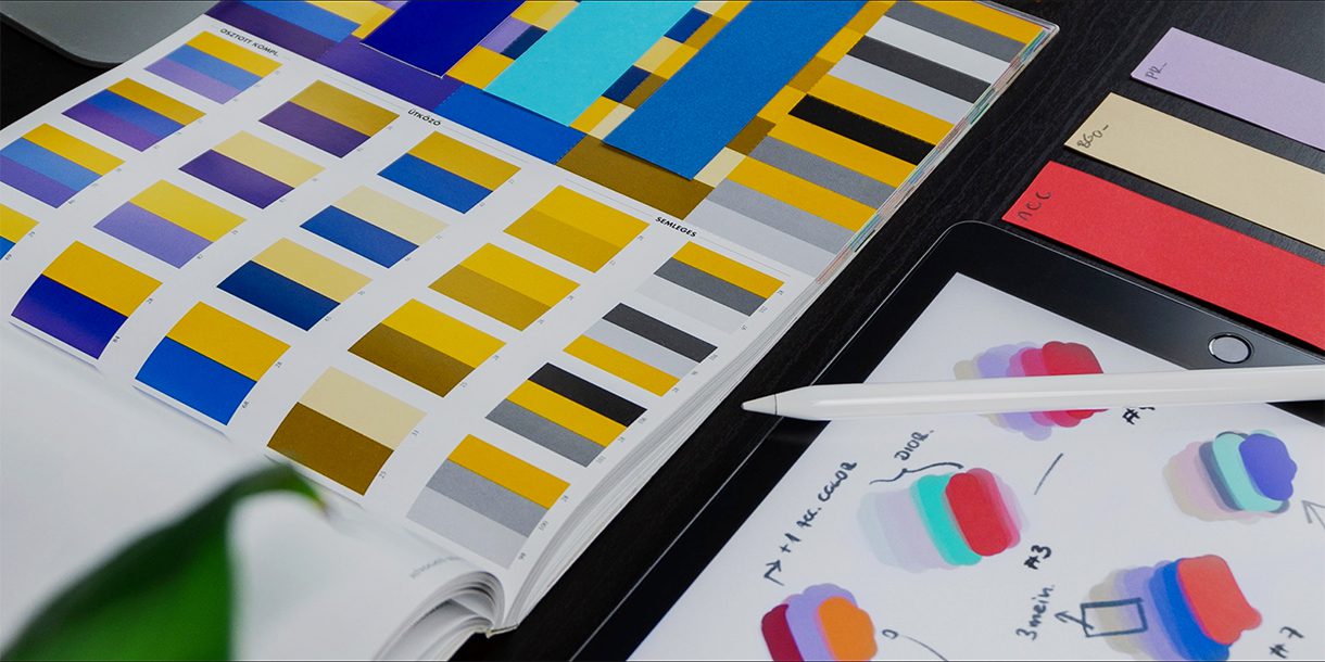 Close-up of a page of a color palette book, paper color swatches, and a stylus resting on a digital tablet.