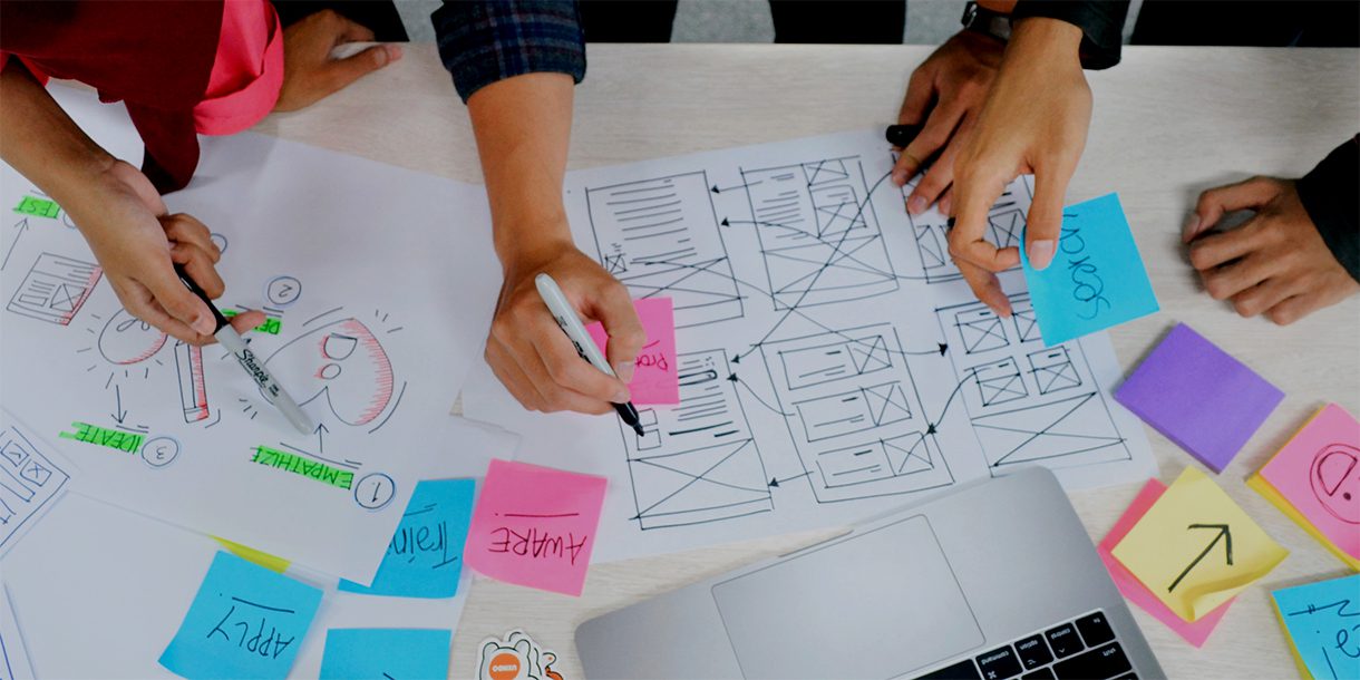 Close-up of a work surface covered with concept drawings, a website wireframe, an open laptop, and sticky notes.