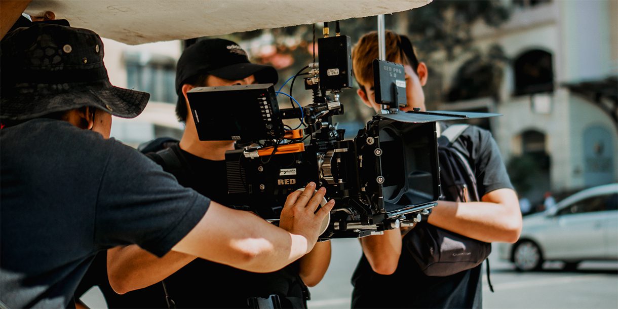 A director, a camera operator holding a large digital video camera, and an assistant shooting content on a city street.
